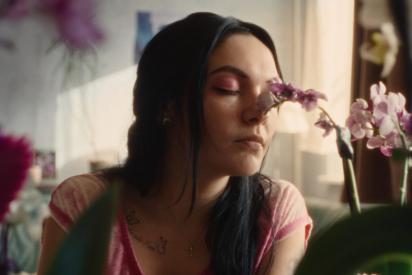 Szene aus dem Film „Vena“: Jenny (gespielt von Emma Nova) mit pinkem Lidschatten und pinkem Shirt sowie langen schwarzen Haaren riecht mit geschlossenen Augen an einer Orchidee.