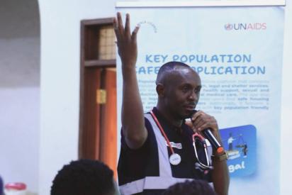 Ein Mann mit Stethoskop um den Hals spricht in ein Mikro, die andere Hand erhoben, dahinter ein Banner von UNAIDS