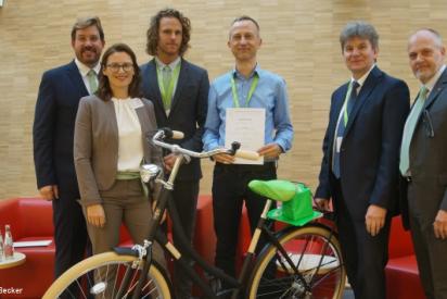 Sechs Personen nebeneinander, in der Mitte ein Mann mit Urkunde in der Hand, davor ein Fahrrad