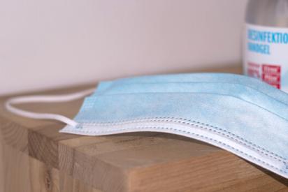 Eine medizinische Maske liegt auf einem Holztisch, im Hintergrund eine Flasche mit Desinfektionsmittel