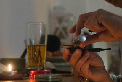 Auf einem Tisch befinden sich verschiedene Drogen: Ein Glas Bier, ein Glas Wein, eine Tüte mit Cannabis, zudem ein Sniefröhrchen, ein brennendes Teelicht und ein Mörser. Davor sitzt eine Person, die sich etwas in eine Pfeife füllt.