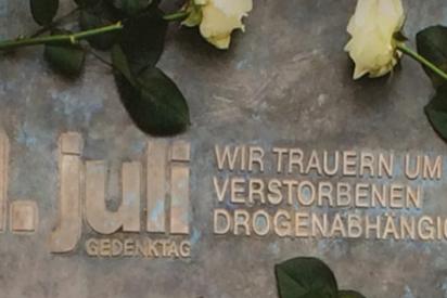 Zu sehen ist ein Gedenkstein mit der Aufschrift: 21. Juli, Gedenktag, wir trauern um die verstorbenen Drogenabhängigen. Frische weiße Rosen umrahmen die Aufschrift.