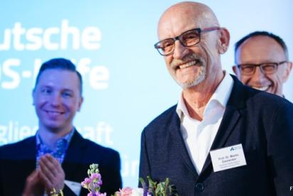 Im Vordergrund: ein lächelnder älterer Mann mit Glatze und dunkler Brille im Anzug, im Hintergrund zwei weitere lächelnde, applaudierende Männer im Anzug