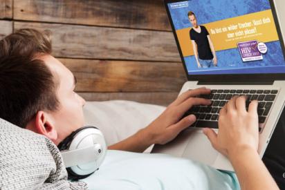 Junger Mann mit Kopfhörern liegt auf einem Bett und surft mit einem Laptop im Internet. Auf dem Laptop-Bildschirm ist die Website www.macht-doch-jeder.de zu sehen.