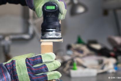 Person trägt Arbeitshandschuhe und schleift mit einem Werkzeug ein Holzteil